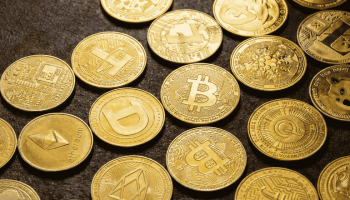 A high-definition, close-up view of various physical golden cryptocurrency coins—including Bitcoin, Ethereum, Dogecoin, and Litecoin—scattered across a dark, textured surface.